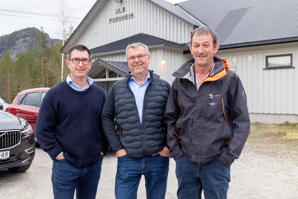 Tre smilende menn står ved siden av hverandre foran en bygning.